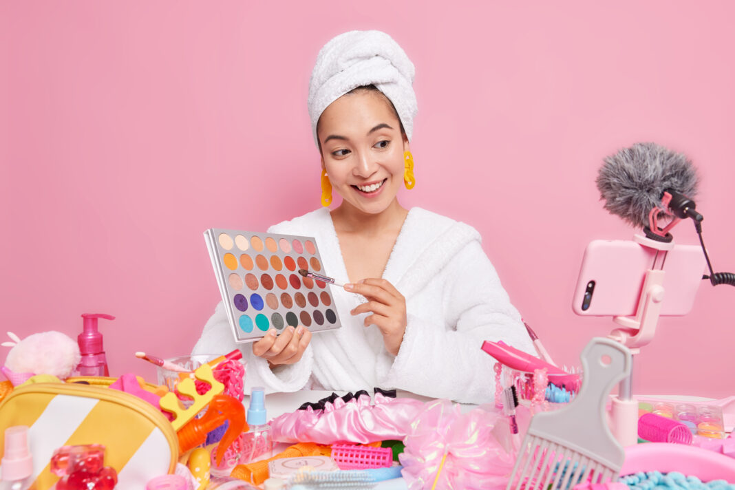 Beautiful pleased Asian woman holds eye shadow palette going to do make up talks with followers wears white bathrobe towel over head focused at smartphone webcam records cosmetic product review