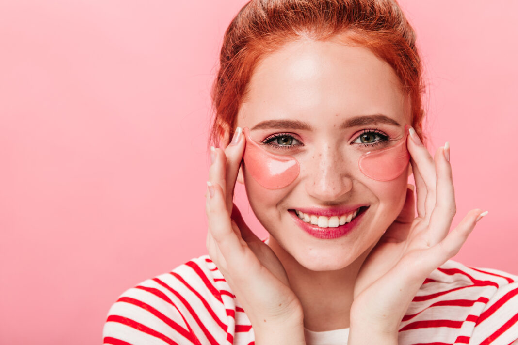 Front view of cheerful pretty woman with eye patches. Happy ginger girl doing skincare treatment and looking at camera